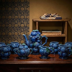 May include: A blue ceramic tea set comprising a teapot, sugar bowl, creamer, and mugs. The set has a fruit design and is displayed on a wooden surface. The background features a patterned wall and a small shelf.