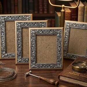 May include: A set of decorative picture frames with gold-coloured edges and detailed silver floral patterns. The frames are displayed on a wooden surface, with a magnifying glass, a small hammer, and a decorative metal band.