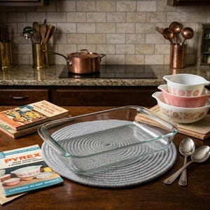 May include: A kitchen scene featuring vintage Pyrex cookware, cookbooks, and serving spoons. A clear glass baking dish rests on a grey placemat. Pink and white patterned bowls are stacked nearby, with a copper pot and stove in the background.