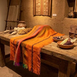 May include: A rustic wooden table displays a vibrant orange and multi-colored woven textile. The textile is draped over the table, with a fringed edge. Bowls of dried goods and a leather satchel are also on the table, with a backdrop of woven wall hangings.