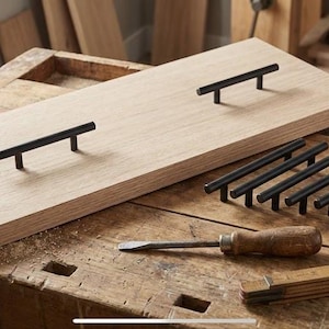 Könnte beinhalten: Ein helles Holzbrett mit drei schwarzen Metallstangengriffen. Mehrere zusätzliche schwarze Stangengriffe befinden sich neben dem Brett. Ein Meißel und ein Zollstock liegen ebenfalls auf der Werkbank.