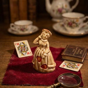 Peut inclure: Figurine en céramique d'une femme en robe avec des découpes en forme de cœur, cartes à jouer, loupe et petit livre. La figurine est sur une surface en velours rouge, avec tasses à thé et théière en arrière-plan. Le livre indique "Wade Pottery: English Heritage 1970".