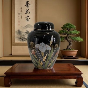 May include: A black ceramic jar with a lid, featuring a floral design with grey, gold, and green accents. The jar is displayed on a dark wooden table in a room with Japanese-style decor.