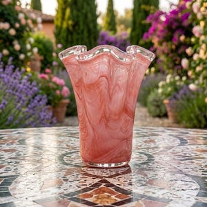 May include: A pink and white marbled glass vase with a wavy rim. The vase is displayed on a mosaic table. The background shows a garden with various flowers and trees, creating a natural setting.
