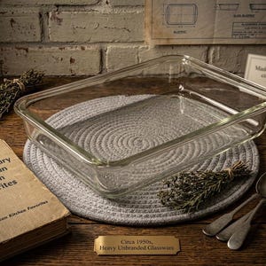 May include: A rectangular glass baking dish is displayed on a grey circular mat. The image includes a vintage cookbook, antique spoons, and text reading "Made in America, circa 1950s". The scene evokes a mid-century kitchen aesthetic, with the dish as the central element.