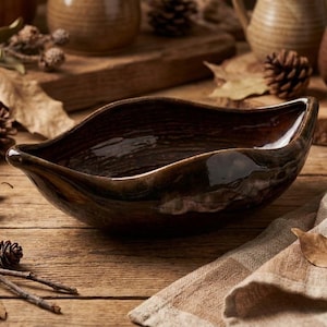 May include: A dark brown, leaf-shaped ceramic bowl with a glossy finish. The bowl is set on a wooden surface, surrounded by pine cones, leaves, and other ceramic pieces. The bowl's interior reflects light.