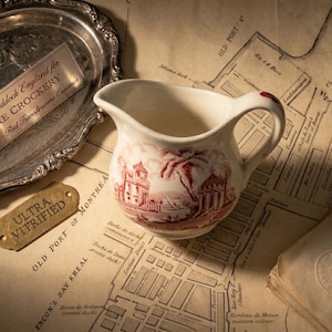Peut inclure: Un pot à crème blanc vintage avec un motif rouge de transfert d'un bâtiment et d'un paysage. Le pot à crème est posé sur une carte avec un plateau argenté et une étiquette métallique qui dit "ULTRA VITRIFIED". Le pot à crème a une anse et un bec.