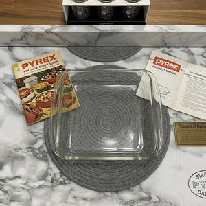 Puede incluir: Una fuente para hornear Pyrex cuadrada y transparente con esquinas redondeadas, junto con un libro de cocina Pyrex vintage y un manual del producto. La imagen también incluye una pequeña placa de latón y un salvamanteles gris. El logotipo de Pyrex y la fecha 1915 son visibles.