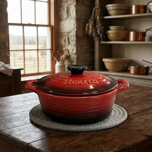 Puede incluir: Una cazuela de cerámica roja con tapa y asas negras, sobre un salvamanteles gris. La palabra "Hearth" está grabada en la tapa. El plato está sobre una mesa de madera rústica en una cocina.