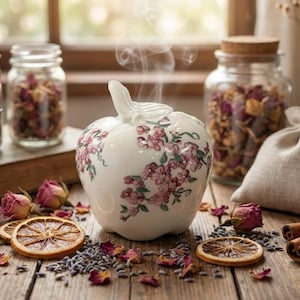 Puede incluir: Un difusor de aceites esenciales con forma de manzana de cerámica blanca con diseños florales rosas. El vapor se eleva desde la parte superior. Capullos de rosa secos, lavanda y rodajas de naranja están esparcidos alrededor del difusor.