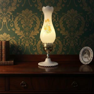 May include: A vintage-style table lamp with a white, scalloped base and a frosted glass shade adorned with yellow floral designs. The lamp sits on a dark wooden surface, alongside a stack of books and a framed portrait, against a green floral wallpaper.