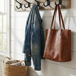 Könnte beinhalten: Ein rustikaler Holz-Kleiderhaken mit vier schwarzen Metallhaken, an dem eine Jeansjacke und eine braune Ledertasche hängen. Ein geflochtener Korb mit gerollten Textilien steht auf einer Holzbank.