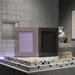 Peut inclure: Deux cadres photo rectangulaires avec un motif de grille métallique, l'un rose pâle et l'autre noir. Une petite plaque dorée devant affiche le texte : VINTAGE POSTMODERN WIRE GRID FRAMES (1980s). Les cadres sont posés sur une surface en terrazzo.