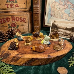 May include: A collection of miniature ceramic figurines of various animals, including a beaver, duck, fish, bison, and deer, displayed on a rustic wooden slice. The figurines are detailed with brown, tan, and cream colors. Vintage tea boxes and a map are in the background.