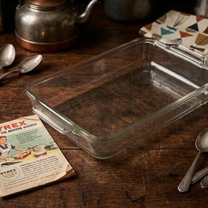 May include: A clear rectangular Pyrex baking dish with handles, on a wooden surface. Vintage spoons and a Pyrex advert are nearby. A patterned cloth and metal kettle are in the background. The advert reads "Pyrex Ware for the Modern Kitchen."