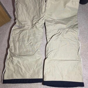 May include: Tan snow pants with white trim and black cuffs. The pants have a quilted texture and the brand name "Converse" is visible on the upper left leg. The pants are laid out on a neutral-colored carpet.