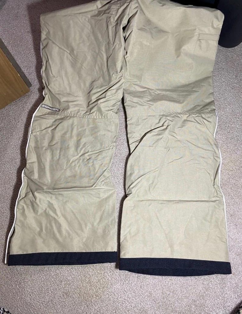 May include: Tan snow pants with white trim and black cuffs. The pants have a quilted texture and the brand name "Converse" is visible on the upper left leg. The pants are laid out on a neutral-colored carpet.