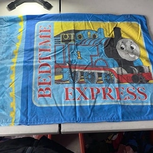 May include: Blue pillowcase featuring a cartoon image of Thomas the Tank Engine. The pillowcase has the words "BEDTIME EXPRESS" in red letters. The background is yellow and blue.