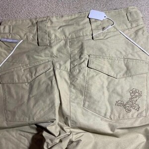 May include: Khaki-colored pants with two back pockets and a decorative floral design on the right pocket. White piping runs along the sides. A blank tag is attached to the waistband.
