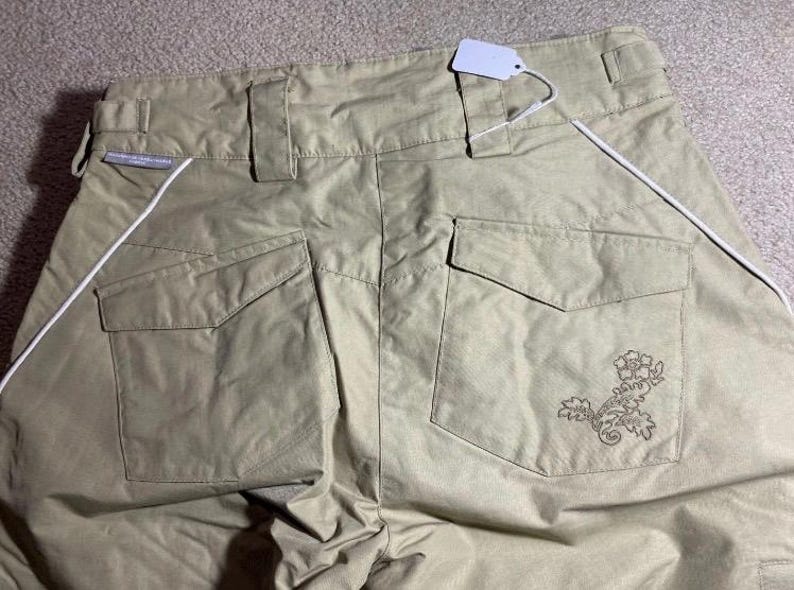 May include: Khaki-colored pants with two back pockets and a decorative floral design on the right pocket. White piping runs along the sides. A blank tag is attached to the waistband.