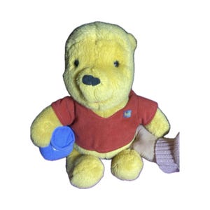 May include: A yellow Winnie the Pooh plush toy wearing a red shirt and holding a blue bucket. The plush has a black nose and small eyes. The shirt has a small "Pooh" patch.