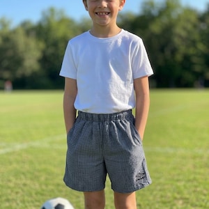 May include: A young person wearing a white t-shirt, black and white checkered shorts, and blue soccer socks stands on a green field. A soccer ball is on the ground. The person is smiling.