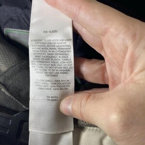 May include: A close-up of a white tag with care instructions and fabric composition details. The tag is held by a hand and is attached to a garment. The text includes washing instructions and fabric content information.