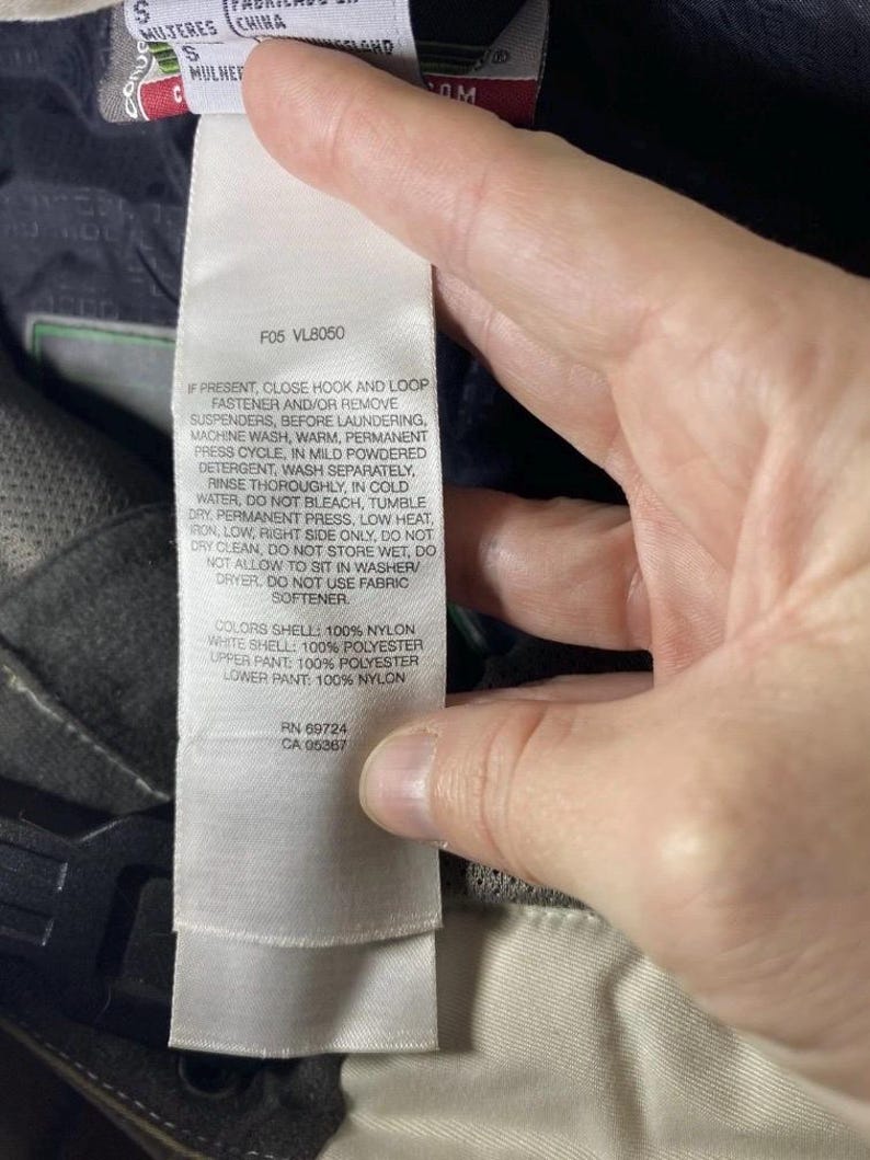 May include: A close-up of a white tag with care instructions and fabric composition details. The tag is held by a hand and is attached to a garment. The text includes washing instructions and fabric content information.
