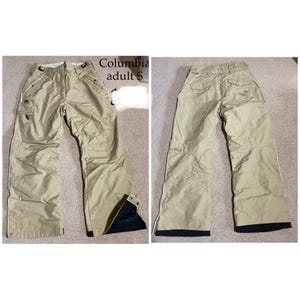 May include: Tan Columbia adult S snow pants. The pants have a straight leg design with white trim along the seams and black accents at the ankles. The front view shows pockets and a zipper closure. The back view shows pockets and a logo.