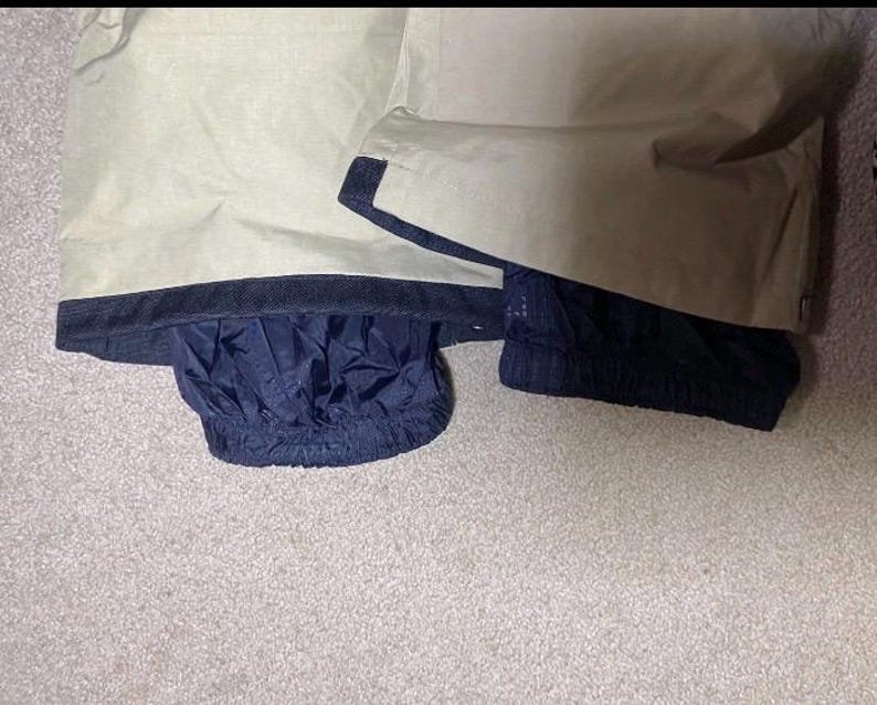 May include: Tan and navy blue snow pants. The pants have a tan outer layer with navy blue accents at the ankles. The ankles have an elasticized cuff. The pants appear to be made of a durable, water-resistant material.