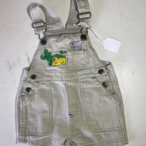 Op de afbeelding: Lichtgrijze baby-overalls met verstelbare bandjes en een voorzak. De zak heeft geborduurde afbeeldingen van een groene alligator, een gele leeuw en een grijze neushoorn. De overalls hebben knoopsluitingen en een blanco label.