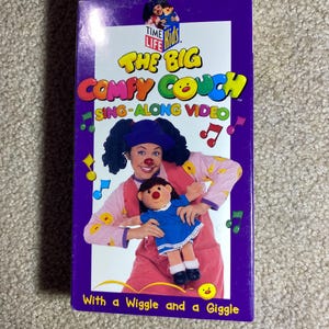 May include: A VHS tape for "The Big Comfy Couch" sing-along video. The cover is purple with the show's title in yellow and green. The image features a woman in clown makeup holding a doll. The text reads "With a Wiggle and a Giggle."