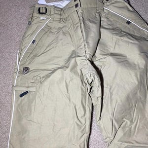 May include: Tan snow pants with white piping along the seams and pockets. The pants have a black logo patch and black accents on the pockets and belt loops. The pants are made of a durable, water-resistant material.