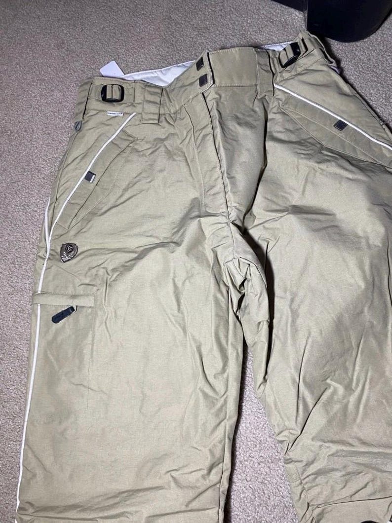May include: Tan snow pants with white piping along the seams and pockets. The pants have a black logo patch and black accents on the pockets and belt loops. The pants are made of a durable, water-resistant material.