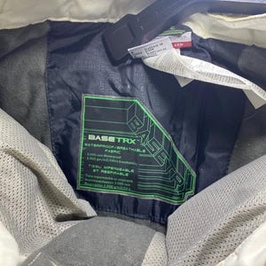 May include: Close-up of a black jacket interior with a green and black label that reads "BASETRX WATERPROOF/BREATHABLE FABRIC". The jacket has a white collar and a black hanger.