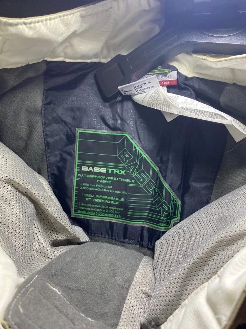 May include: Close-up of a black jacket interior with a green and black label that reads "BASETRX WATERPROOF/BREATHABLE FABRIC". The jacket has a white collar and a black hanger.