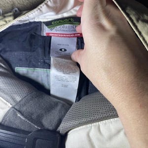 May include: Close-up of the interior of a pair of tan Columbia Convert Sportswear Comp pants. The pants have a black interior with a white tag that reads "convert" and "Columbia Sportswear Comp". The tag also includes the website CONVERTBOARDSWEAR.COM.