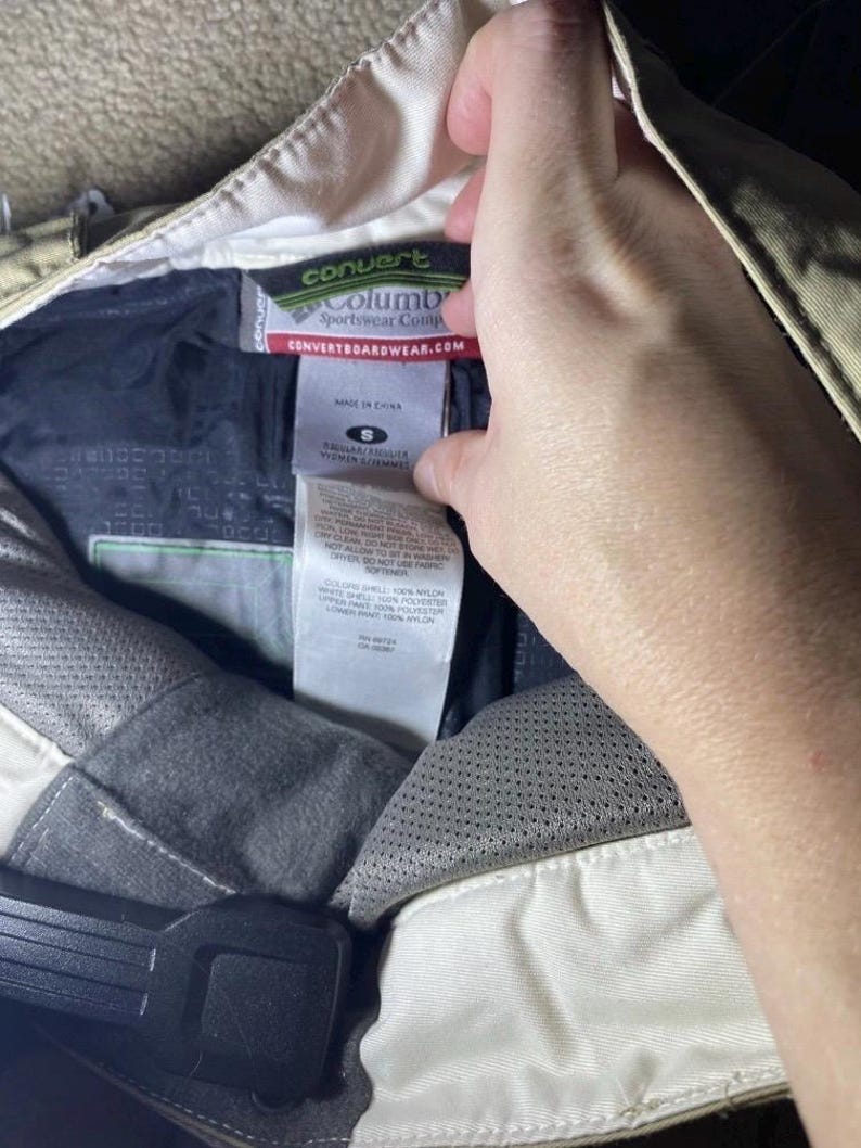 May include: Close-up of the interior of a pair of tan Columbia Convert Sportswear Comp pants. The pants have a black interior with a white tag that reads "convert" and "Columbia Sportswear Comp". The tag also includes the website CONVERTBOARDSWEAR.COM.