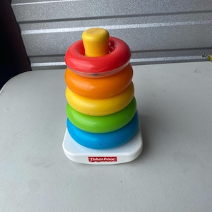 Può includere: Un giocattolo impilabile Fisher-Price con un piolo giallo e cinque anelli colorati. Gli anelli sono rossi, arancioni, gialli, verdi e blu, impilati su una base bianca. Un giocattolo classico per bambini piccoli.
