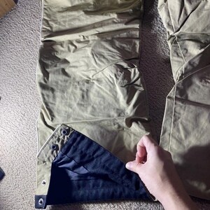 May include: Close-up of olive-green pants with a black interior lining. The pants have a button closure at the ankle and a white trim. The fabric appears to be lightweight and slightly wrinkled.