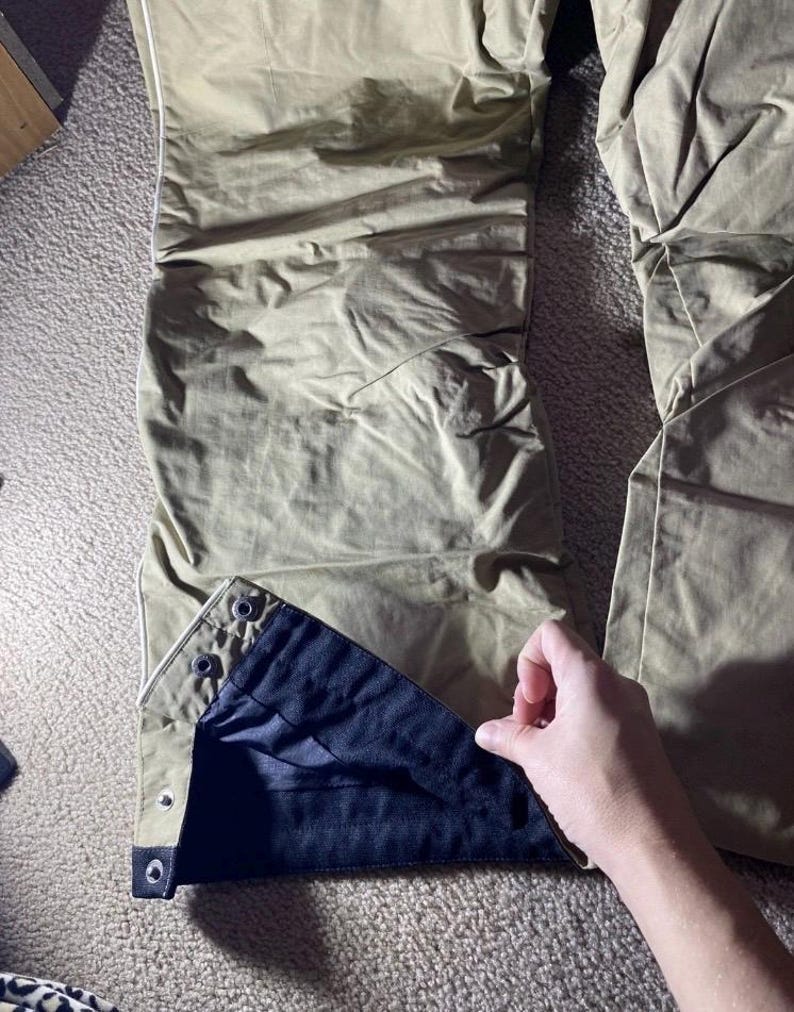 May include: Close-up of olive-green pants with a black interior lining. The pants have a button closure at the ankle and a white trim. The fabric appears to be lightweight and slightly wrinkled.