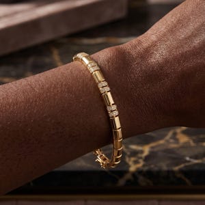 Op de afbeelding: Een gouden armband met rechthoekige schakels en diamantaccenten. De armband wordt om een pols gedragen, wat het ontwerp en het vakmanschap benadrukt. De sluiting is zichtbaar en de algehele uitstraling is luxueus en elegant.