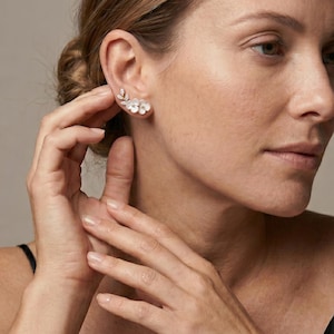 May include: A silver-toned ear climber earring featuring a delicate floral design with three white petals and a small leaf-shaped accent adorned with tiny clear stones. The earring follows the curve of the earlobe, creating a layered botanical look.