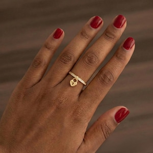 May include: A gold-colored ring with a small padlock charm. The ring band is adorned with small, clear stones. The padlock charm hangs from a bar on the ring. The background is a dark brown color.