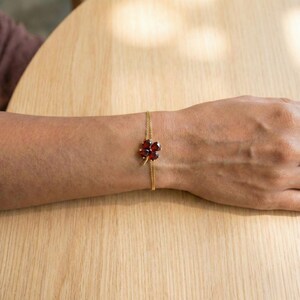May include: A gold-colored bracelet featuring a four-leaf clover design with deep red gemstones. The clover is the focal point, set on a delicate chain. The bracelet is displayed on a person's wrist.