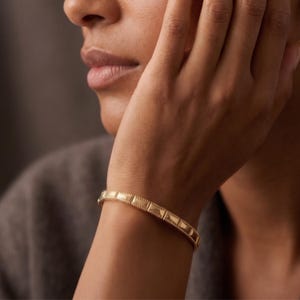 May include: A gold-toned metal bracelet featuring a series of rectangular, slightly raised segments connected by a beaded border design. The bracelet has a polished finish and a structured, link-style appearance that wraps around the wrist.
