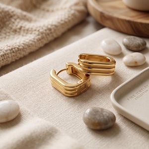 May include: A pair of gold-toned hoop earrings featuring a rectangular shape with three distinct horizontal ridges along the outer surface. The earrings have a hinged closure mechanism and are displayed alongside smooth, rounded river stones.