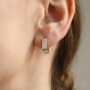 May include: A close-up of a silver earring with a rectangular hoop design. The earring has three parallel bars that form the hoop. The earring is worn on an ear, and the background is blurred.