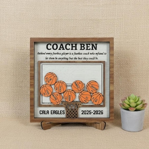 May include: Wooden basketball-themed plaque with the text "COACH BEN" and a motivational quote. The plaque features a basketball court design with several basketballs and the text "CALA EAGLES 2025-2026". A small succulent in a gray pot is on the right.