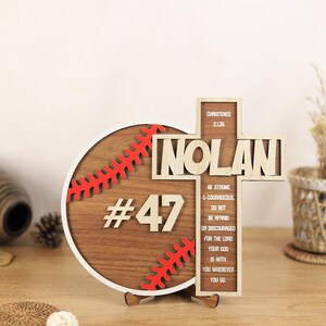 May include: Wooden baseball-themed plaque with the name "NOLAN" and the number "47". The plaque features a baseball design with red stitching and a cross with inspirational text. The text includes "Be strong & courageous" and "Do not be afraid".